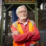 Man in red jumper and orange vest looking at the camera in front of industrial infrastructure