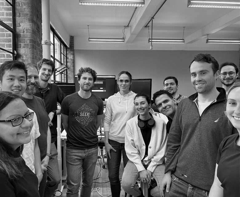 12 people standing in a semi-circle smiling at the camera in an office.