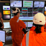 6 adults standing in a cement factory control room. All but one of the adults are in high vis jumpsuits and hard hats. They are standing in front of an array of screens showing graphs and camera footage.