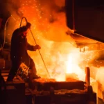 Man in safety jumpsuit holding long stick into vat on molten steel